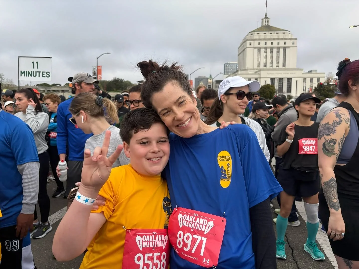 A smiling woman and a boy making a peace sign at the Oakland Marathon, surrounded by other runners and spectators with city buildings in the background.