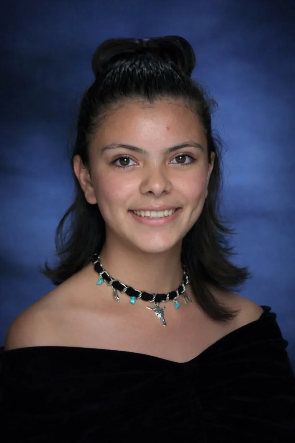 Young woman with dark hair styled in a half-updo, wearing a black off-the-shoulder top, a black and turquoise necklace with charms, smiling against a blue background.