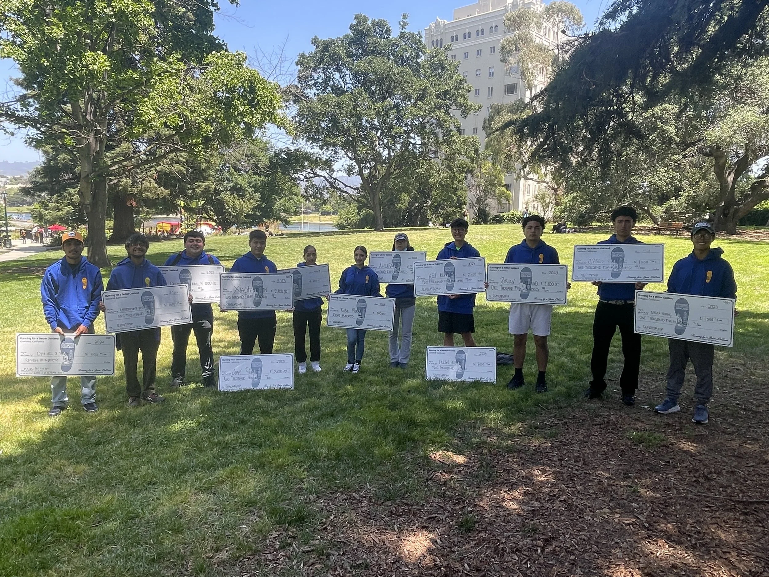 A group of ten young people standing outdoors on grass, holding oversized checks, with trees and a tall white building in the background.