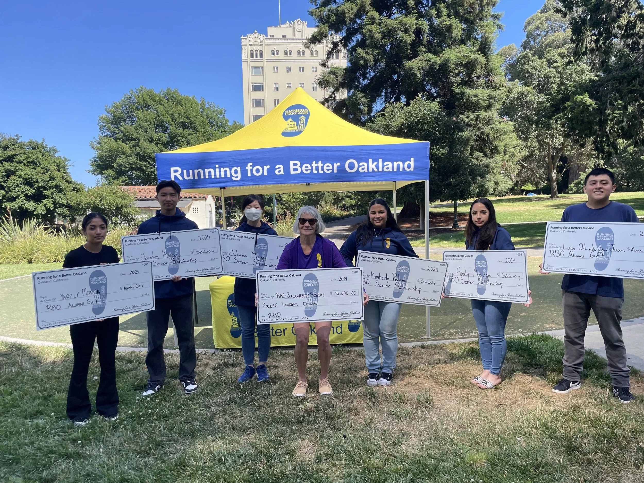 Group of young people holding large checks, standing outdoors in front of a blue and yellow tent with the text 'Running for a Better Oakland', surrounded by trees and a tall building in the background.