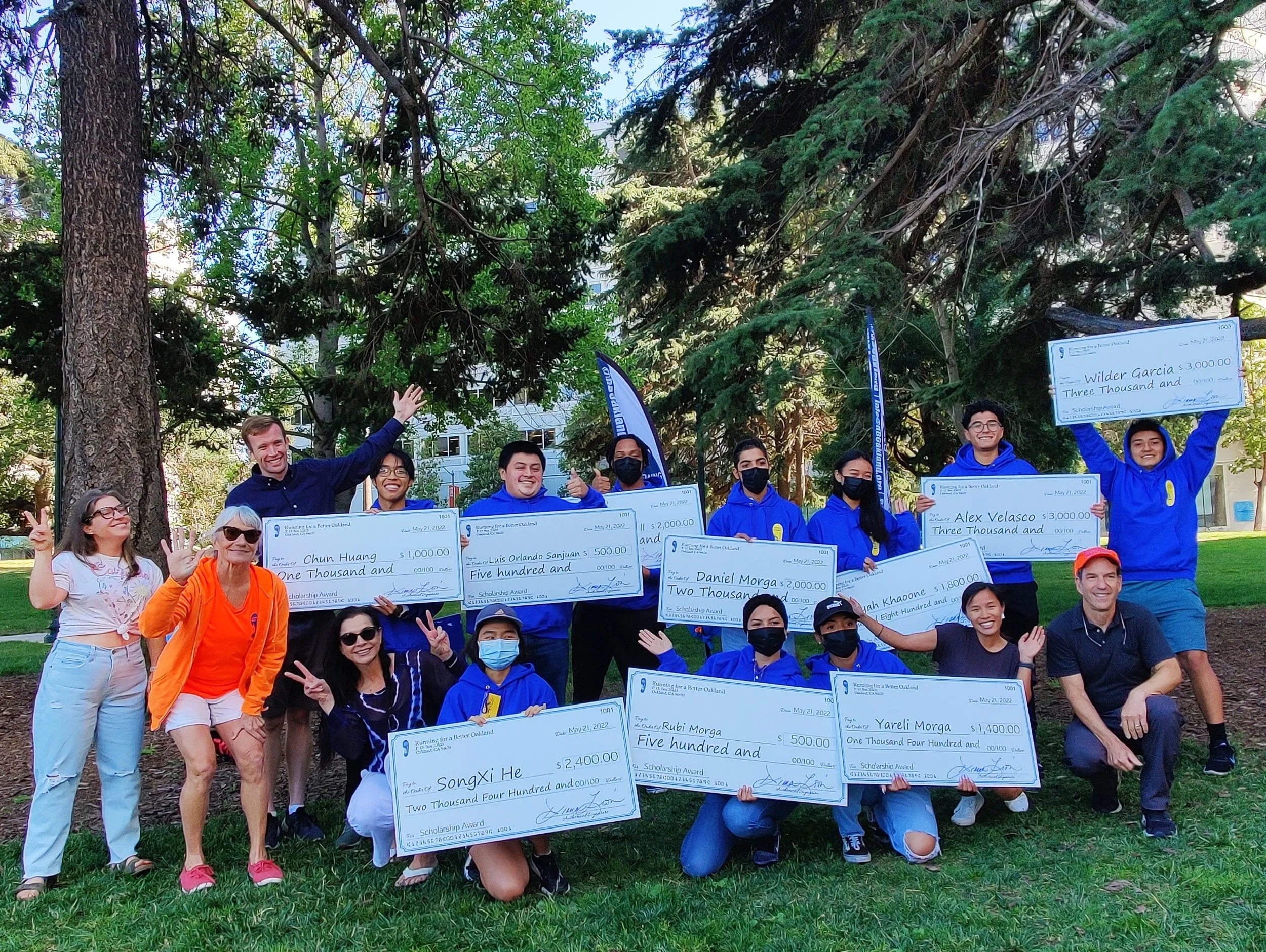 Group of people outdoors holding large symbolic checks, some wearing blue hoodies, others face masks, standing in front of large trees in a park.