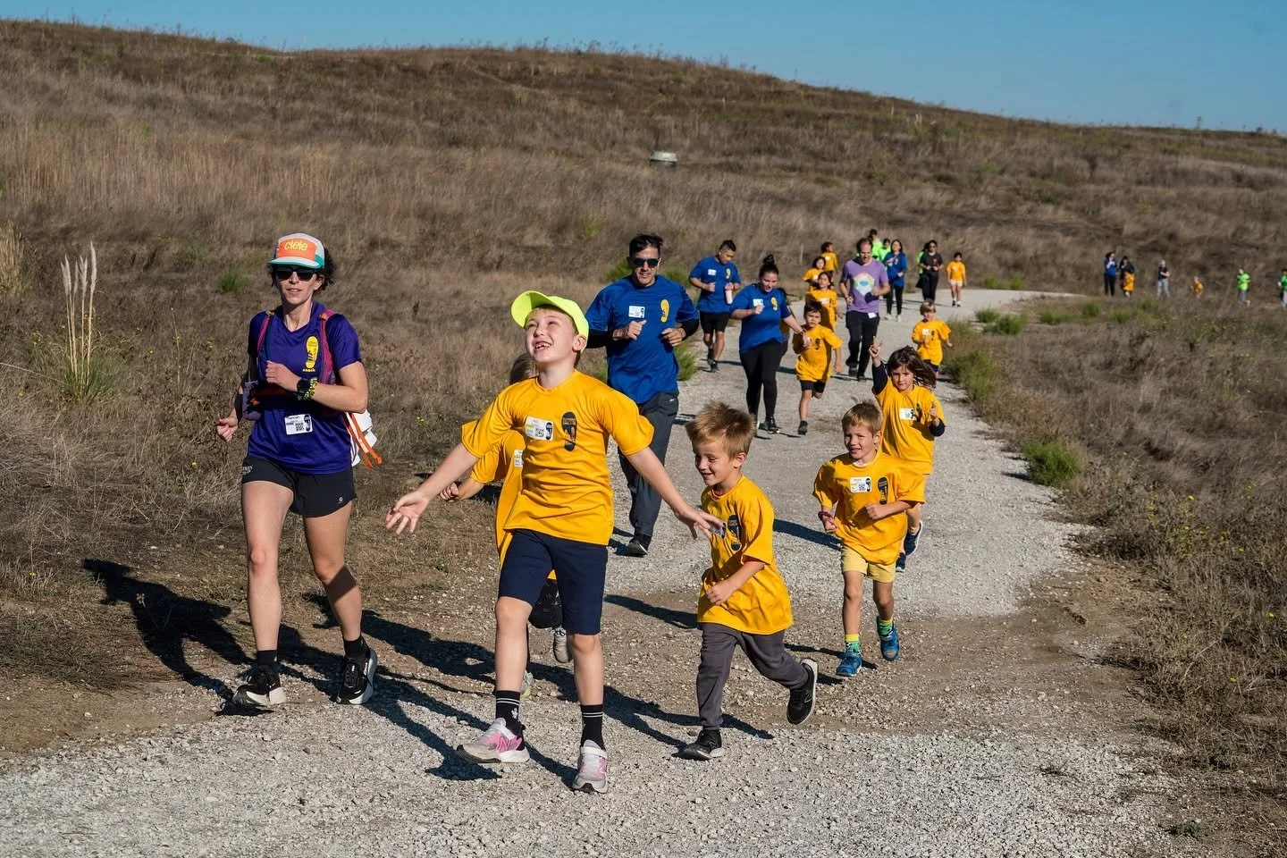 Group of children and adults participating in an outdoor charity run or walk on a dirt trail through a hilly, dry landscape, many wearing yellow shirts, with some in blue shirts, under a clear blue sky.