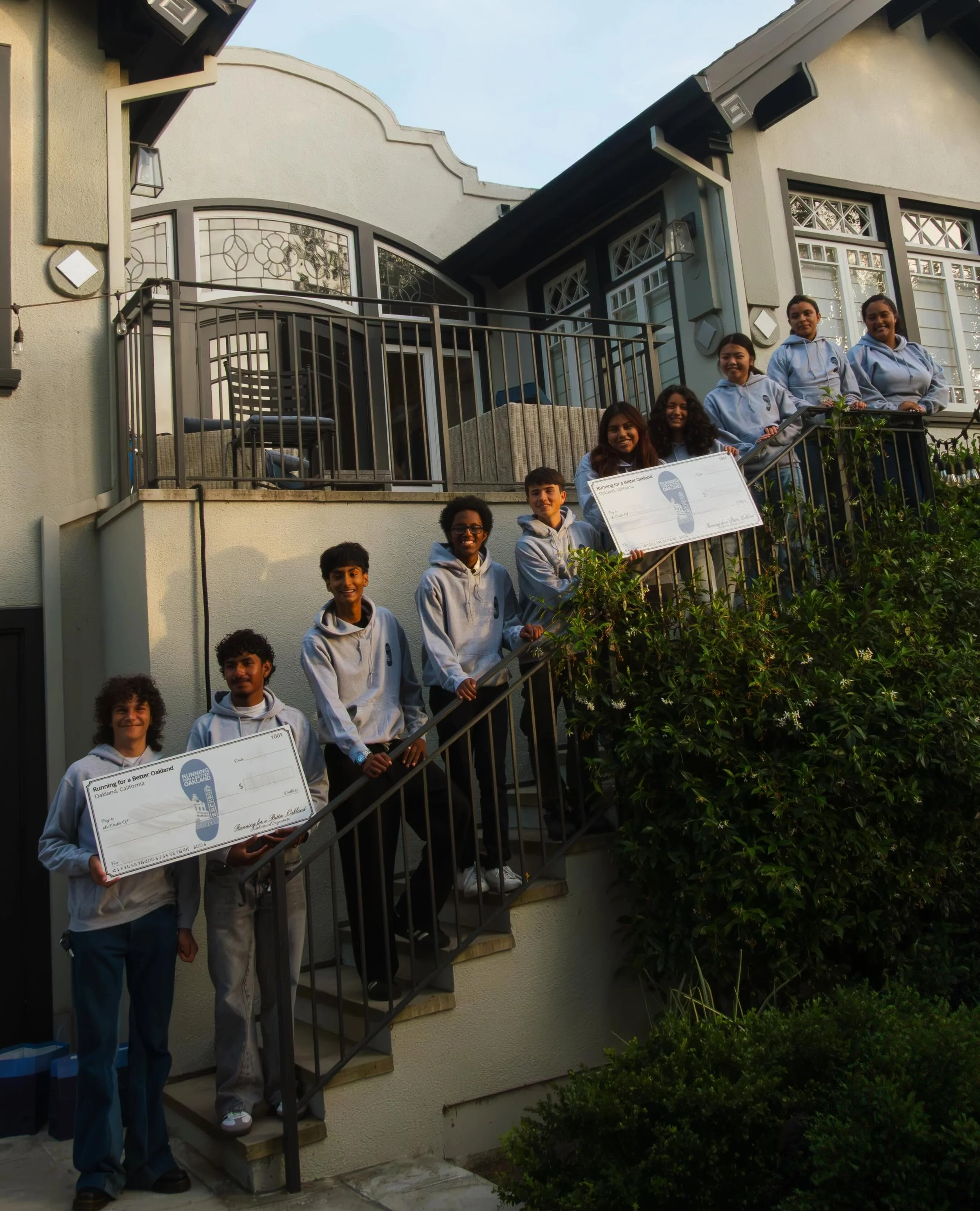 Group of people standing on a staircase outside a house, holding oversized checks and smiling.