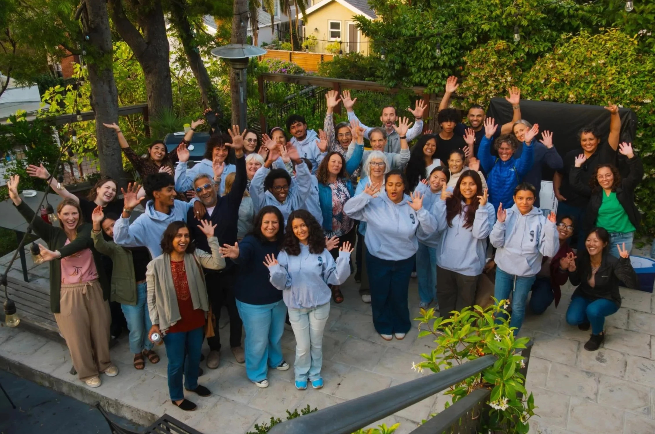 A group of diverse people gathered outdoors, smiling and waving, with trees and a house in the background.