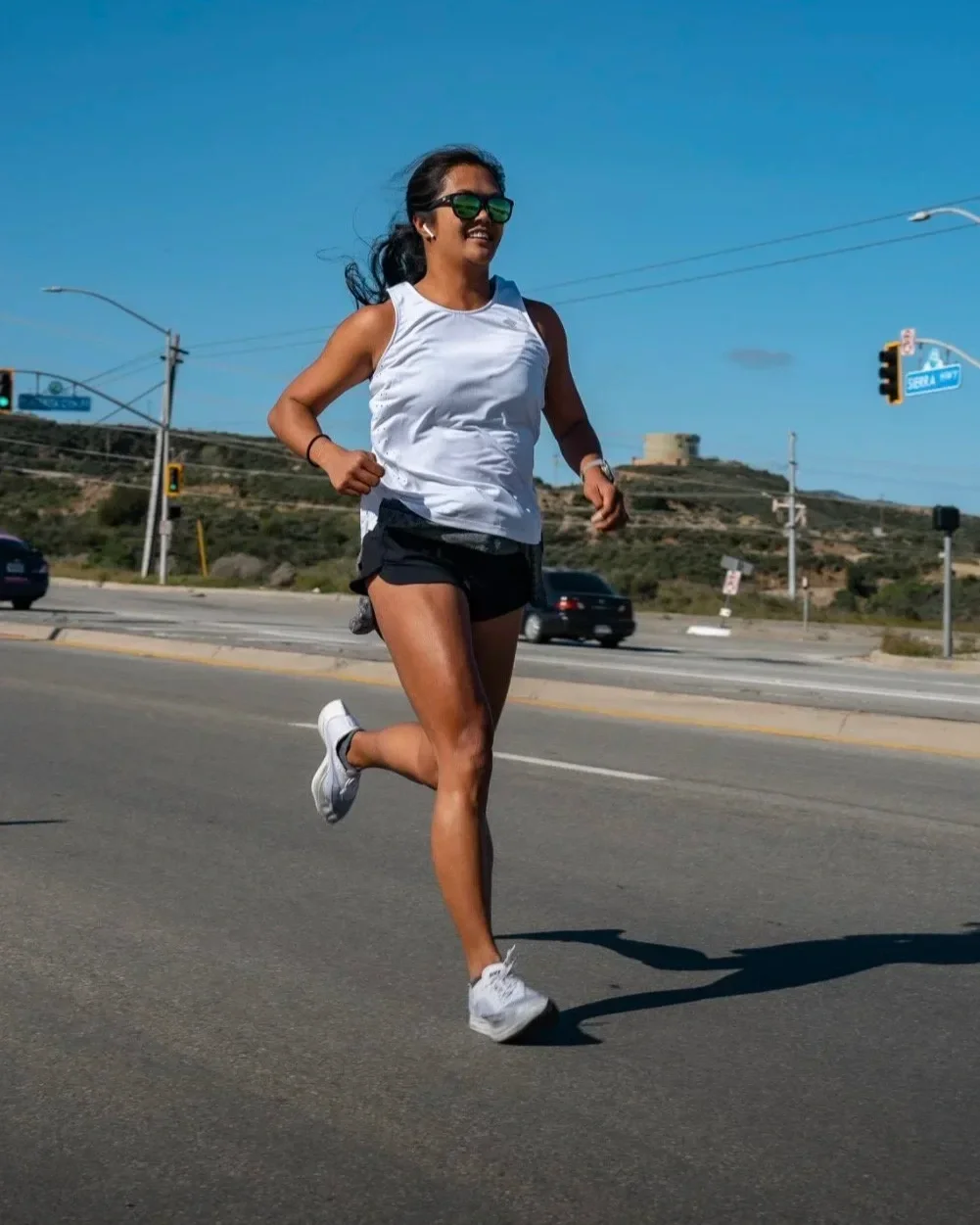 A woman running on a road with a blue sky and hills in the background, wearing sunglasses, a white sleeveless top, black shorts, and white running shoes.