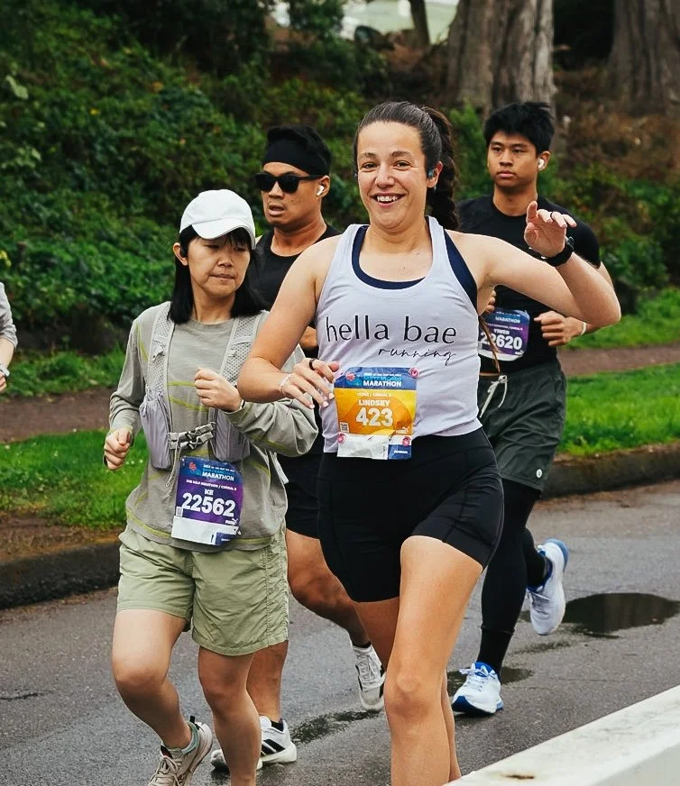 Group of marathon runners, woman smiling and wearing a tank top that says 'hella bae running', participating in a race on a wet trail with greenery and trees in the background.