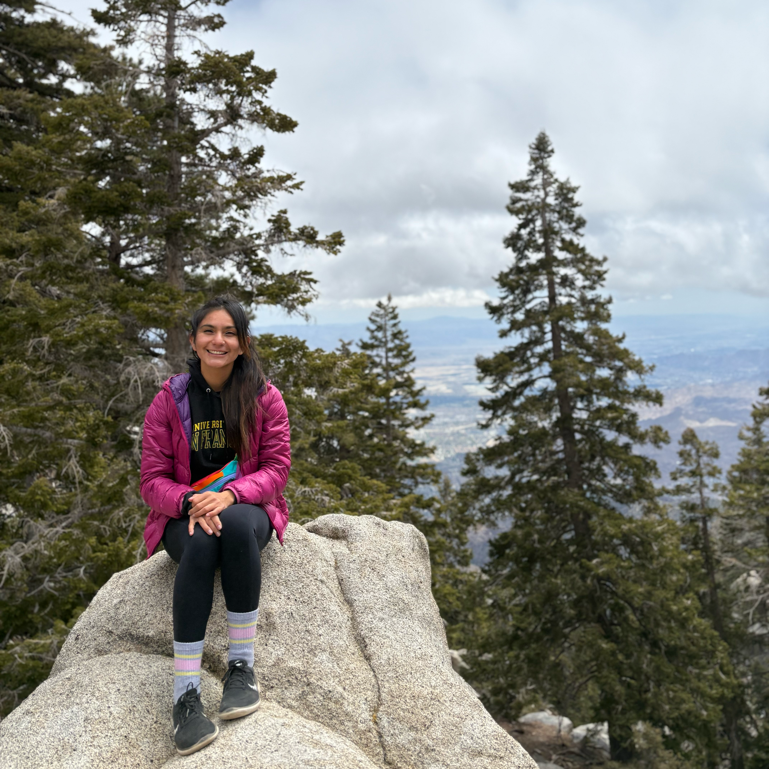 A smiling woman sitting on a large rock among pine trees in a mountainous area, with a view of distant mountains and cloudy sky.