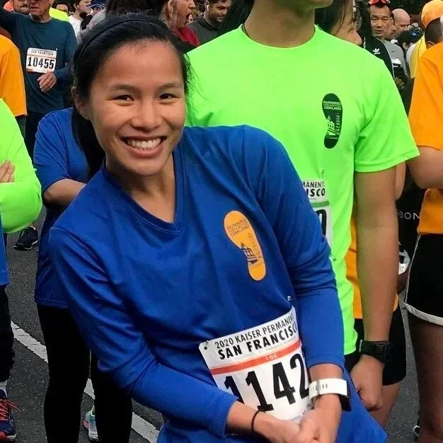 A smiling woman in a blue running shirt with a race bib, surrounded by other runners at a marathon event.