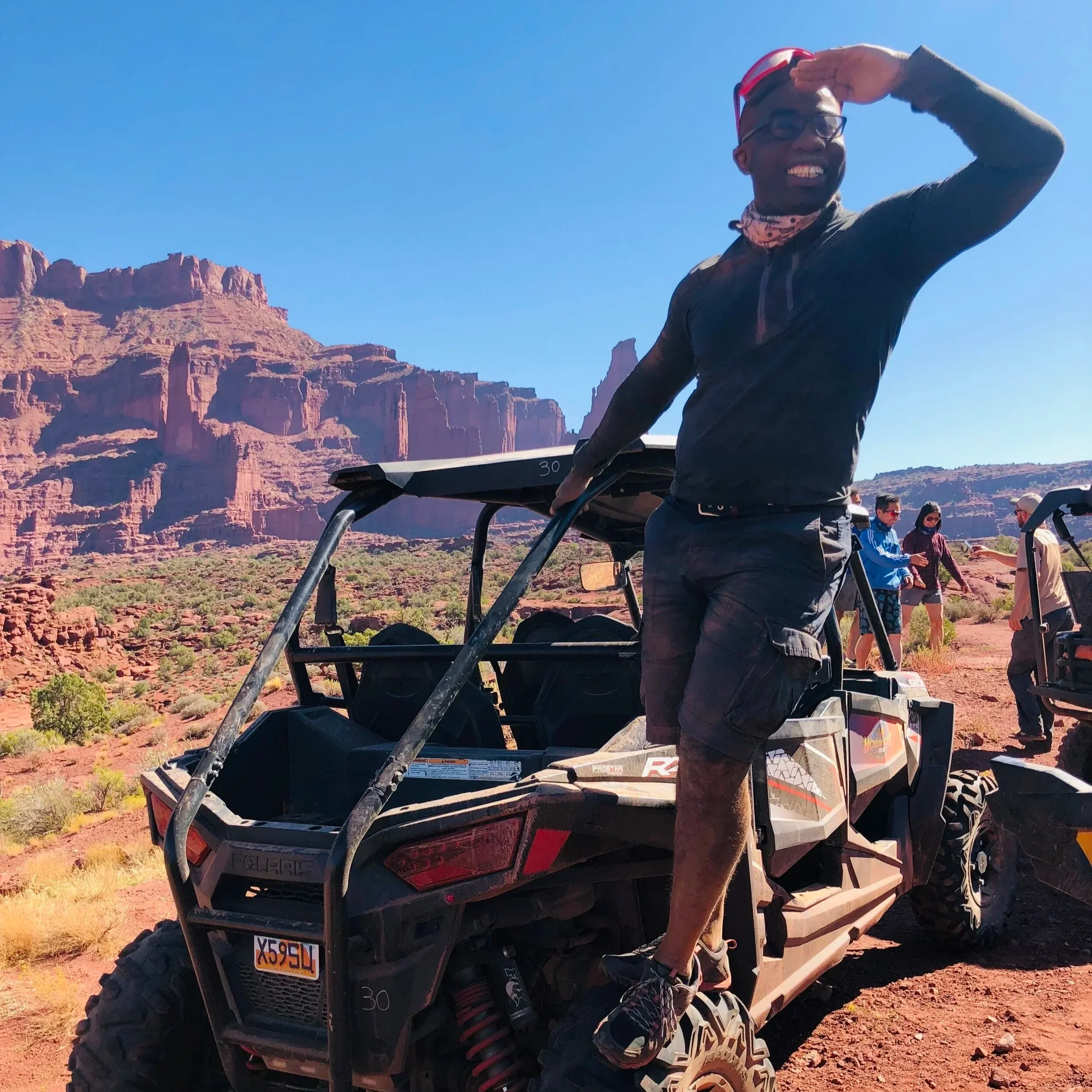 A person standing on a utility vehicle with a desert landscape and rock formations in the background, saluting and smiling under clear blue skies.