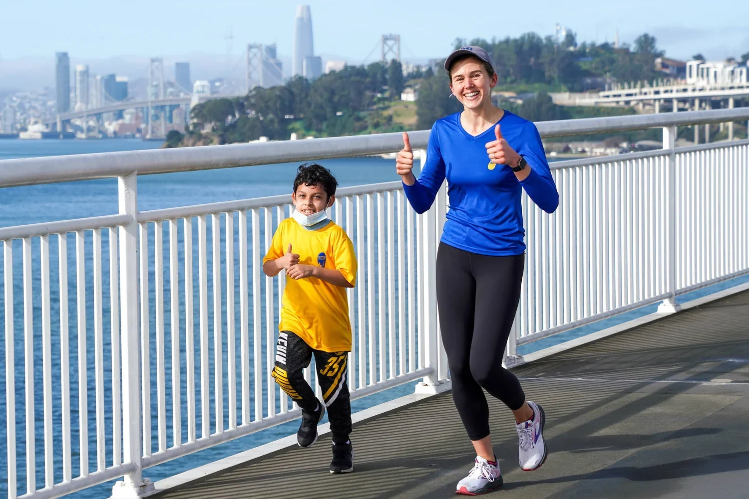 A woman and a young boy jogging on a waterfront promenade with a cityscape and bridge in the background, smiling and giving thumbs up.