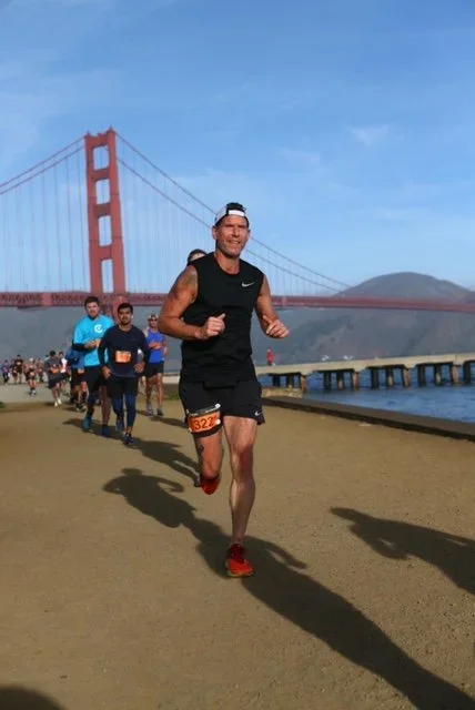 A man running in a marathon near the Golden Gate Bridge with other runners behind him, on a sunny day.