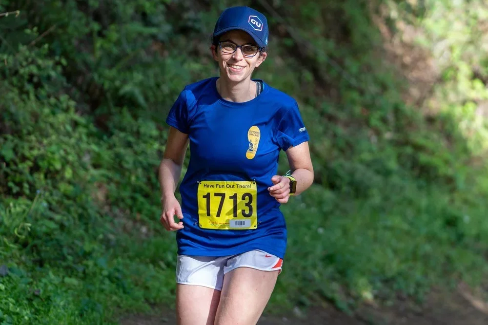 A woman running outdoors on a trail, wearing a blue shirt, white shorts, a dark blue cap, glasses, and a bib number 1713 with the message "Have Fun Out There!"