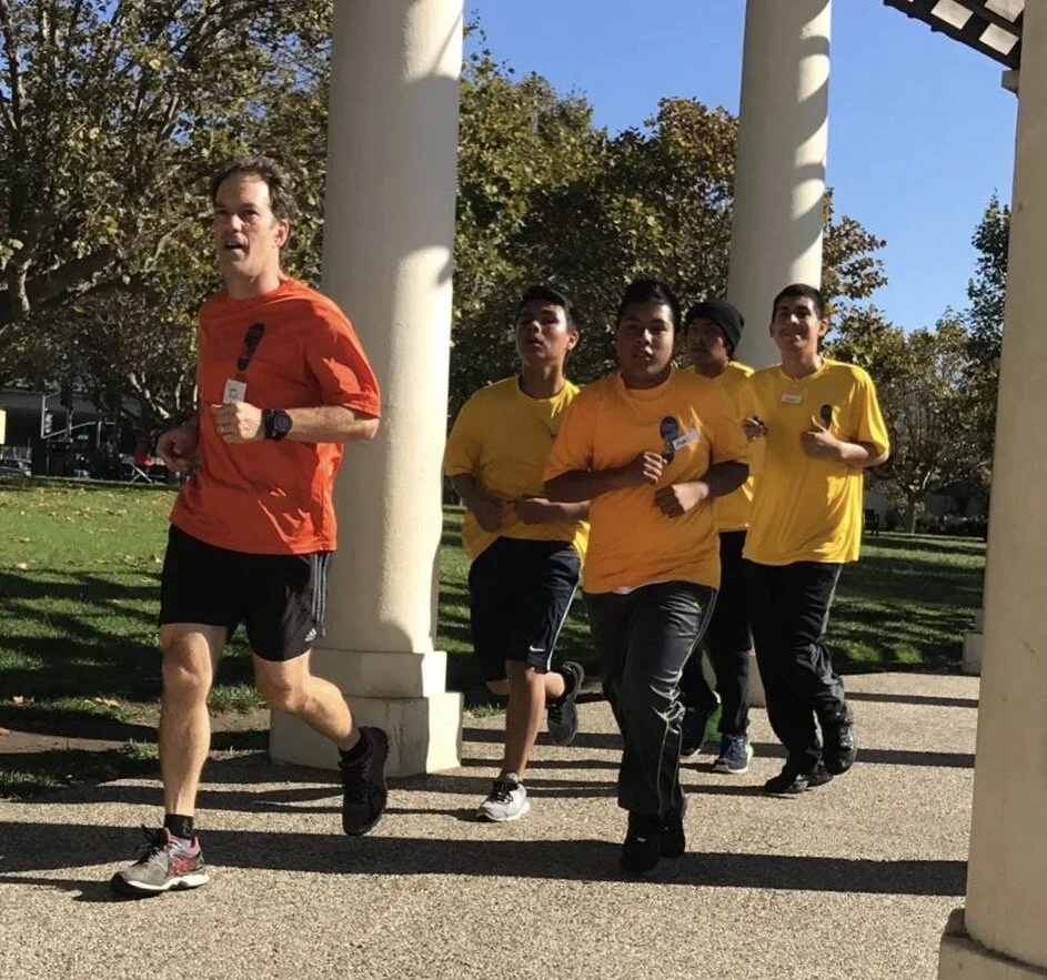 A group of four young men running together outdoors, with one man leading, in a park with trees and large white columns.
