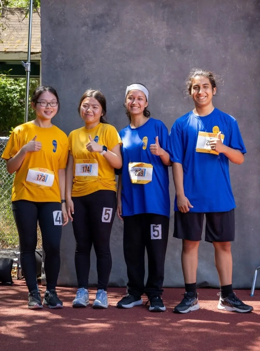 Four diverse young women in yellow and blue athletic shirts with race bibs are standing side by side outdoors, smiling, and giving thumbs-up gestures after a race.