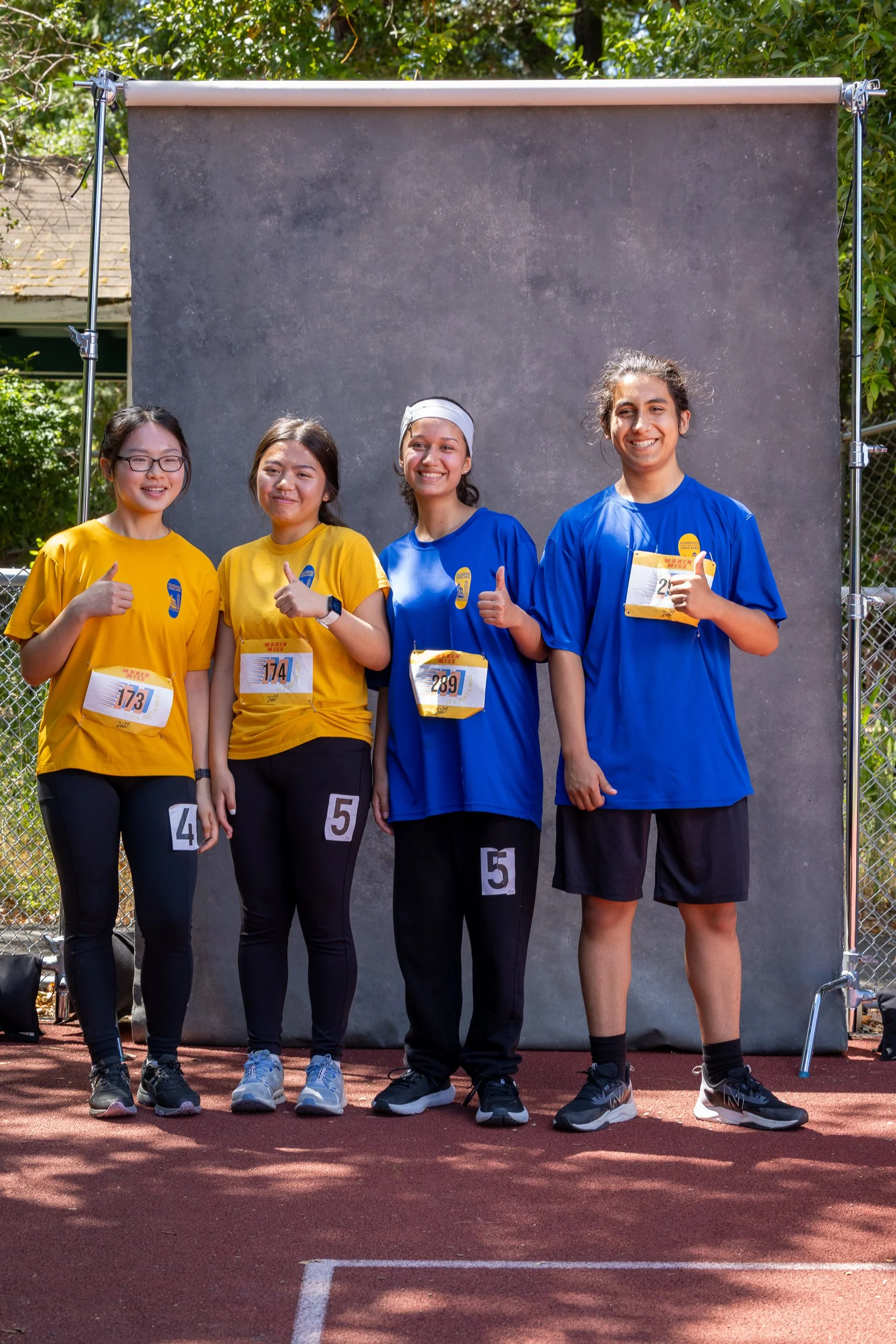 Four young women in athletic shirts and athletic pants standing on a track field in front of a gray backdrop, smiling and giving thumbs up.