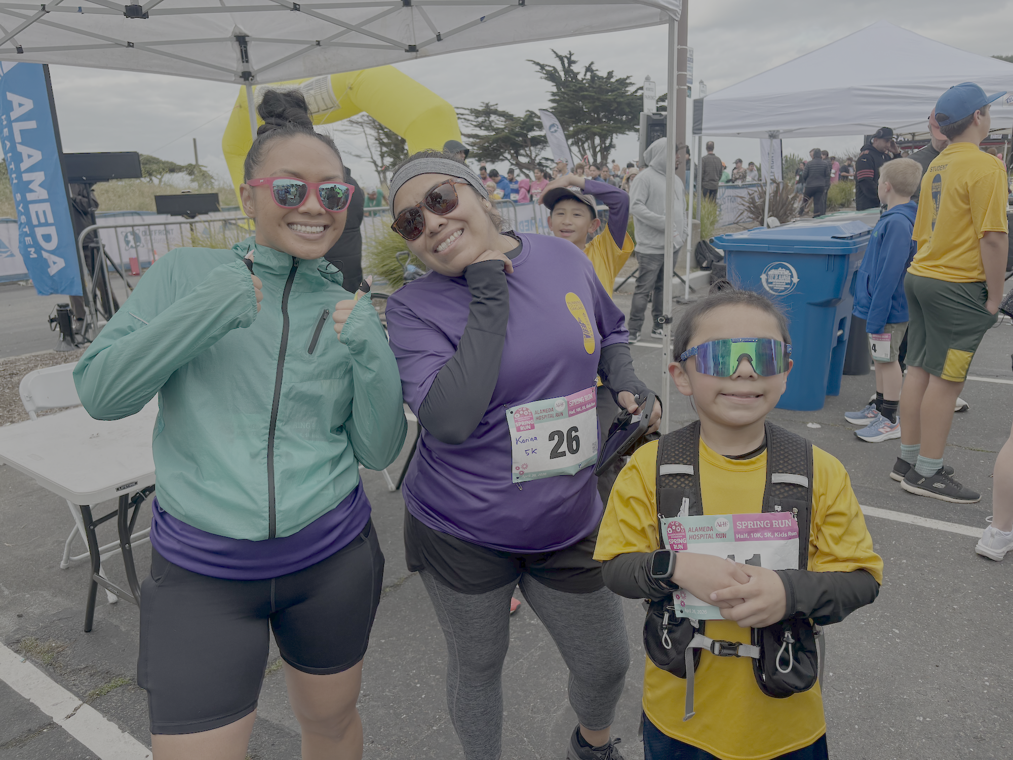 Smiling women and a boy at a running event, standing in front of a starting line with an inflatable arch, Asian women wearing sunglasses and athletic clothing, and a young boy wearing yellow sports clothes and sunglasses