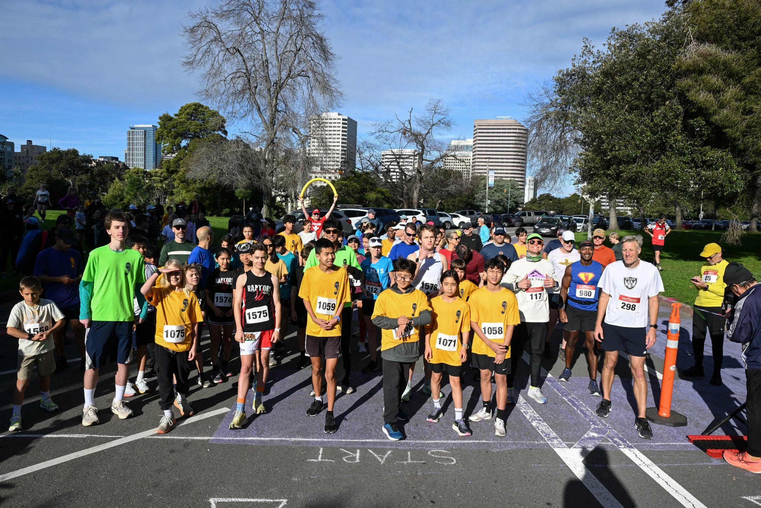Crowd of marathon runners gathered at the starting line in a park with city skyscrapers in the background.