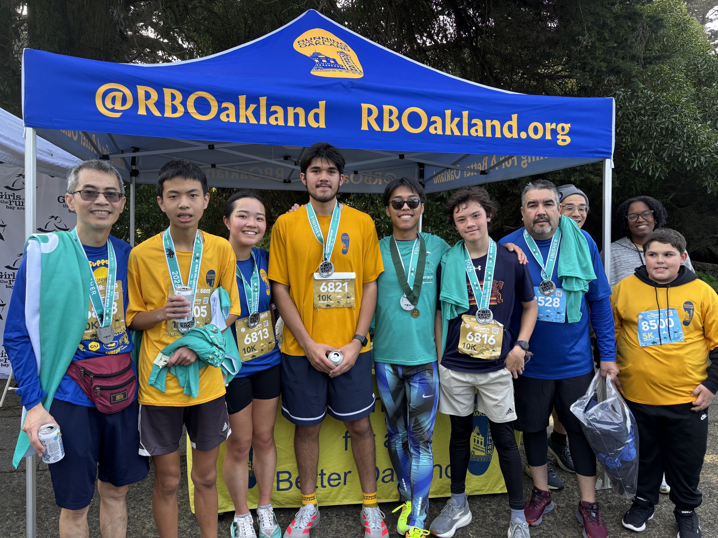 Group of runners wearing medals and race bibs, standing in front of a blue tent with '@RBOakland.org' written on it, at a race event.