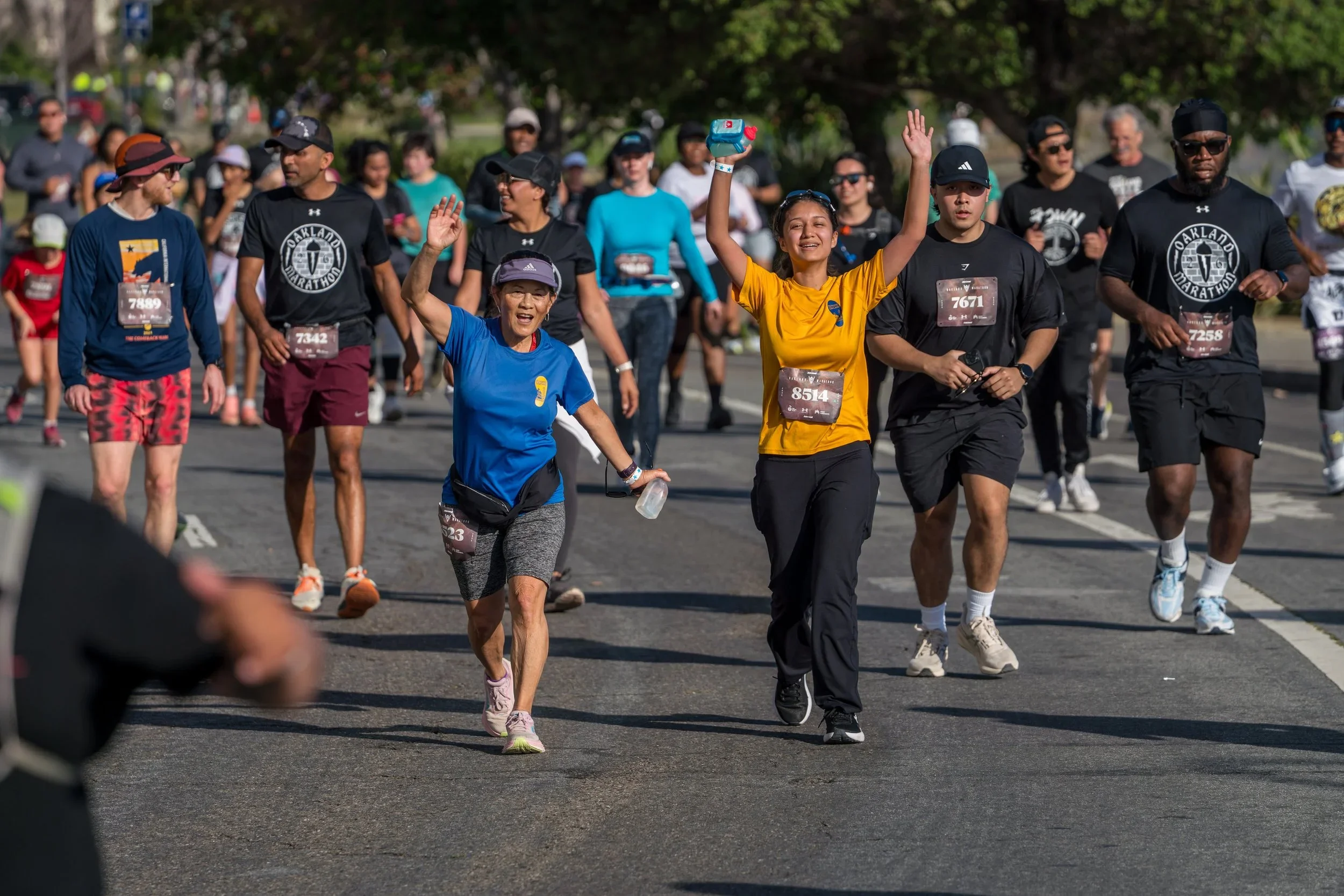 Group of people participating in a marathon or running event, with some smiling and waving, wearing race bibs and athletic clothing, walking or jogging on a tree-lined street.