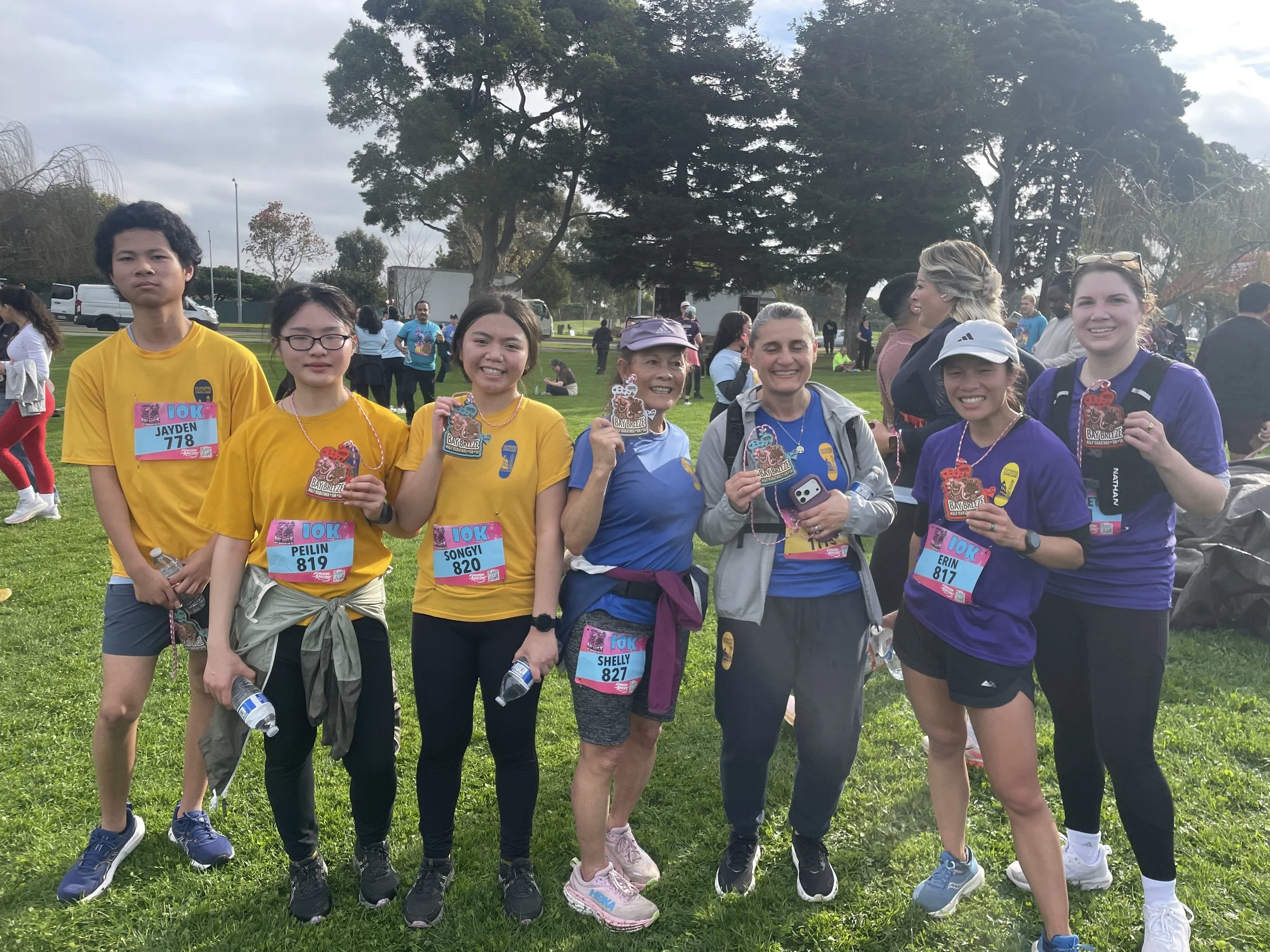 Group of women and young girls at a park running event, holding medals and water bottles, with trees and other participants in the background.