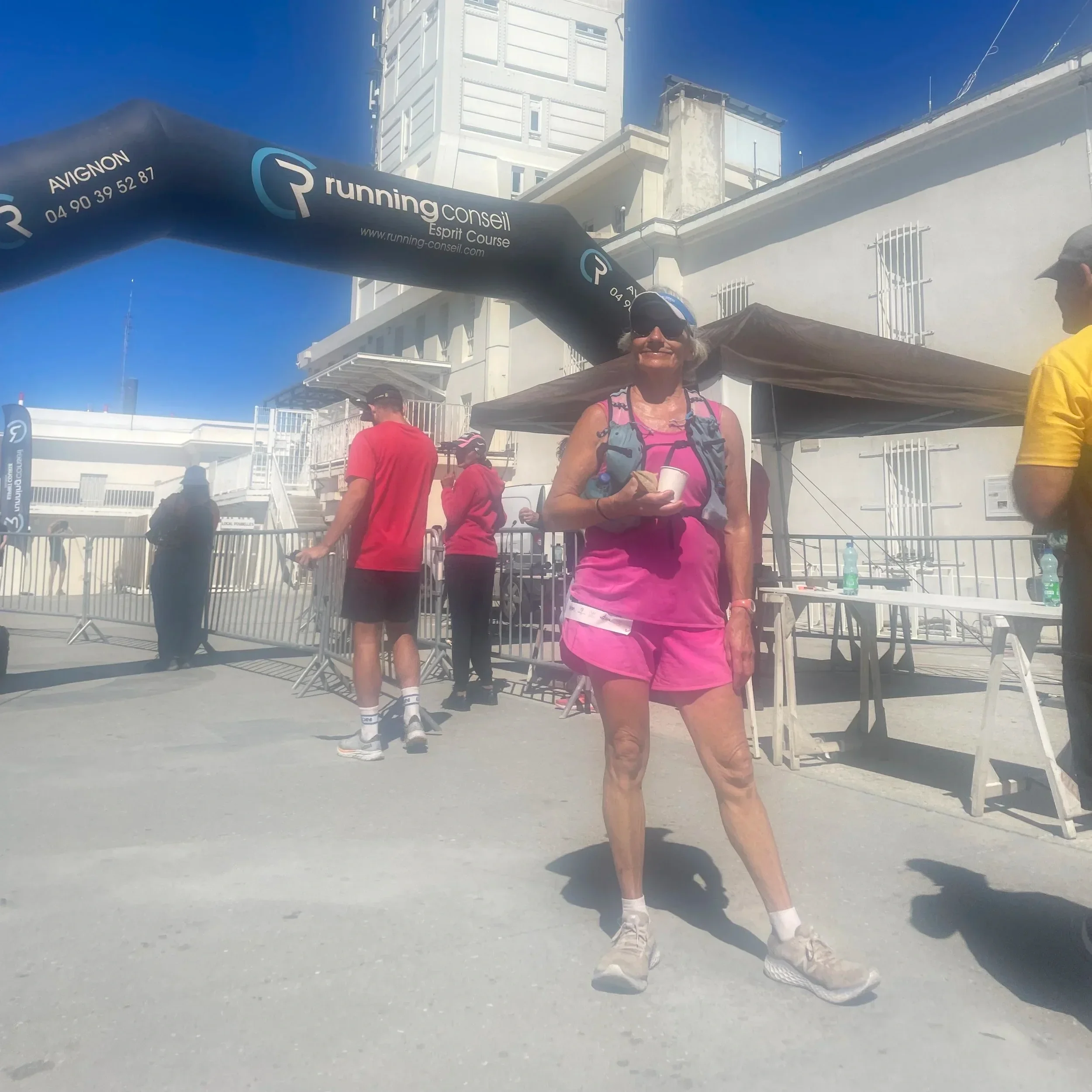 A woman in pink athletic outfit and sneakers stands on a dusty street, holding a cup and smiling. Several people are around her, with a building and a black inflatable arch that has