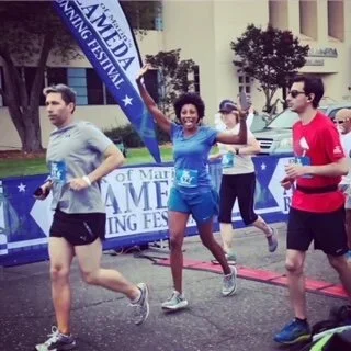 Three runners participate in a marathon, with two men in athletic wear and a woman smiling and waving at the finish line, which has banners and trees in the background.