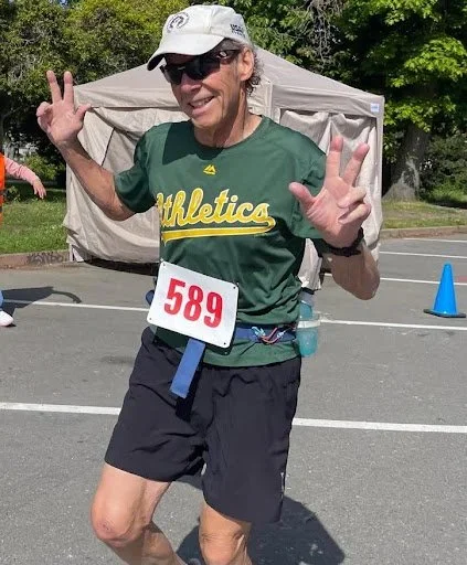 A smiling female runner in a green athletic shirt, black shorts, sunglasses, and a white cap, holding up both hands in a victory gesture at a race event, with a bib number 589.