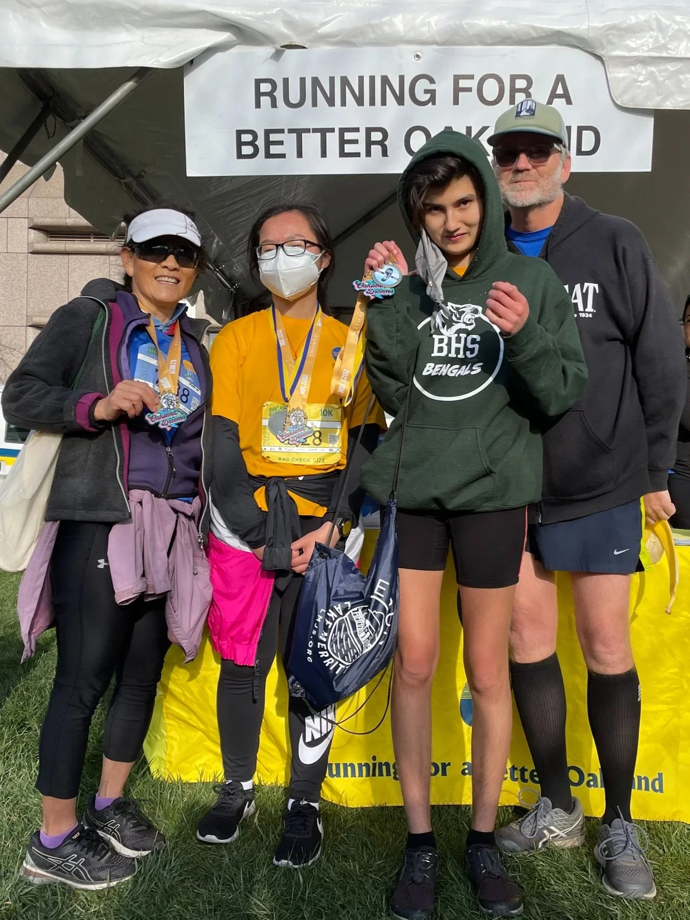 A group of five people, including a young runner with a medal, pose in front of a banner that reads 'Running for a better Oakland.' They are dressed in athletic clothing and standing on grass, smiling and celebrating their participation in a race event.
