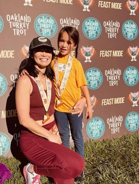 A woman and a young girl are posing for a photo at the Oakland Turkey Trot event, standing in front of a backdrop with event logos. The woman is wearing a maroon outfit and a black cap, and the girl is in a yellow shirt with medals around her neck. They are smiling, with the woman kneeling and the girl standing next to her.