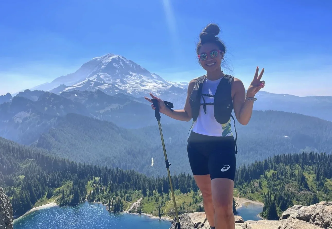 A woman hiking on a mountain trail with a snow-capped mountain in the background, holding hiking poles and making peace signs with both hands, wearing sunglasses, a white shirt, and black shorts.