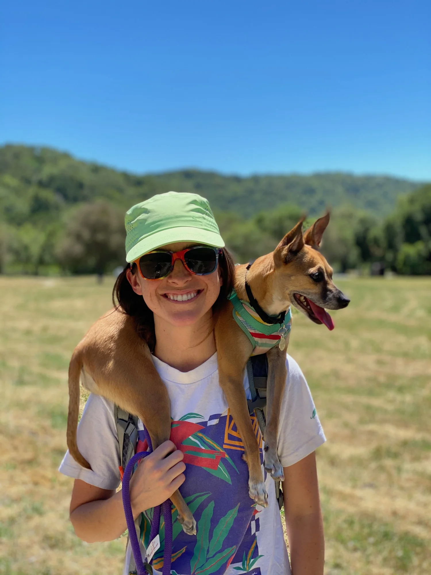 Woman wearing a light green hat and sunglasses, smiling, carrying a tan dog with a colorful harness on her shoulders, in an outdoor field with green hills and a clear blue sky in the background.
