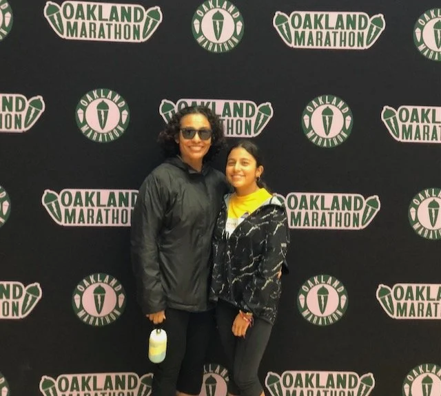 Two women standing in front of an Oakland Marathon branded backdrop, smiling for the camera.
