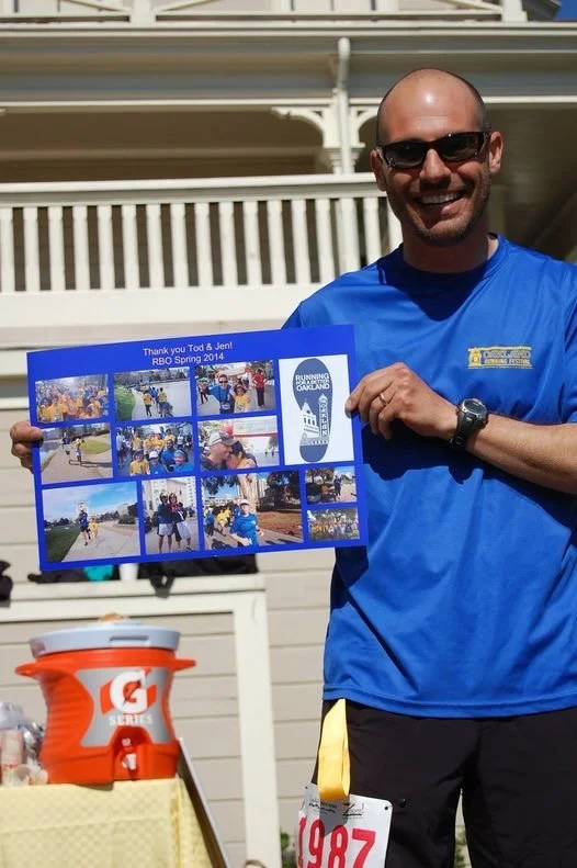 A smiling man wearing sunglasses and a blue athletic shirt holding a large photo collage, standing outdoors near a white building with a balcony, with an orange Gatorade cooler and a yellow container in front of him.
