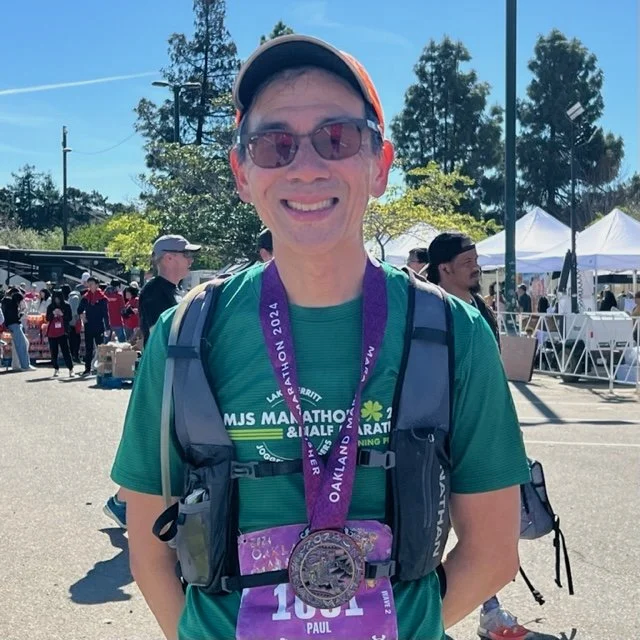 Man wearing sunglasses and a baseball cap, smiling, with a marathon medal around his neck at an outdoor event.