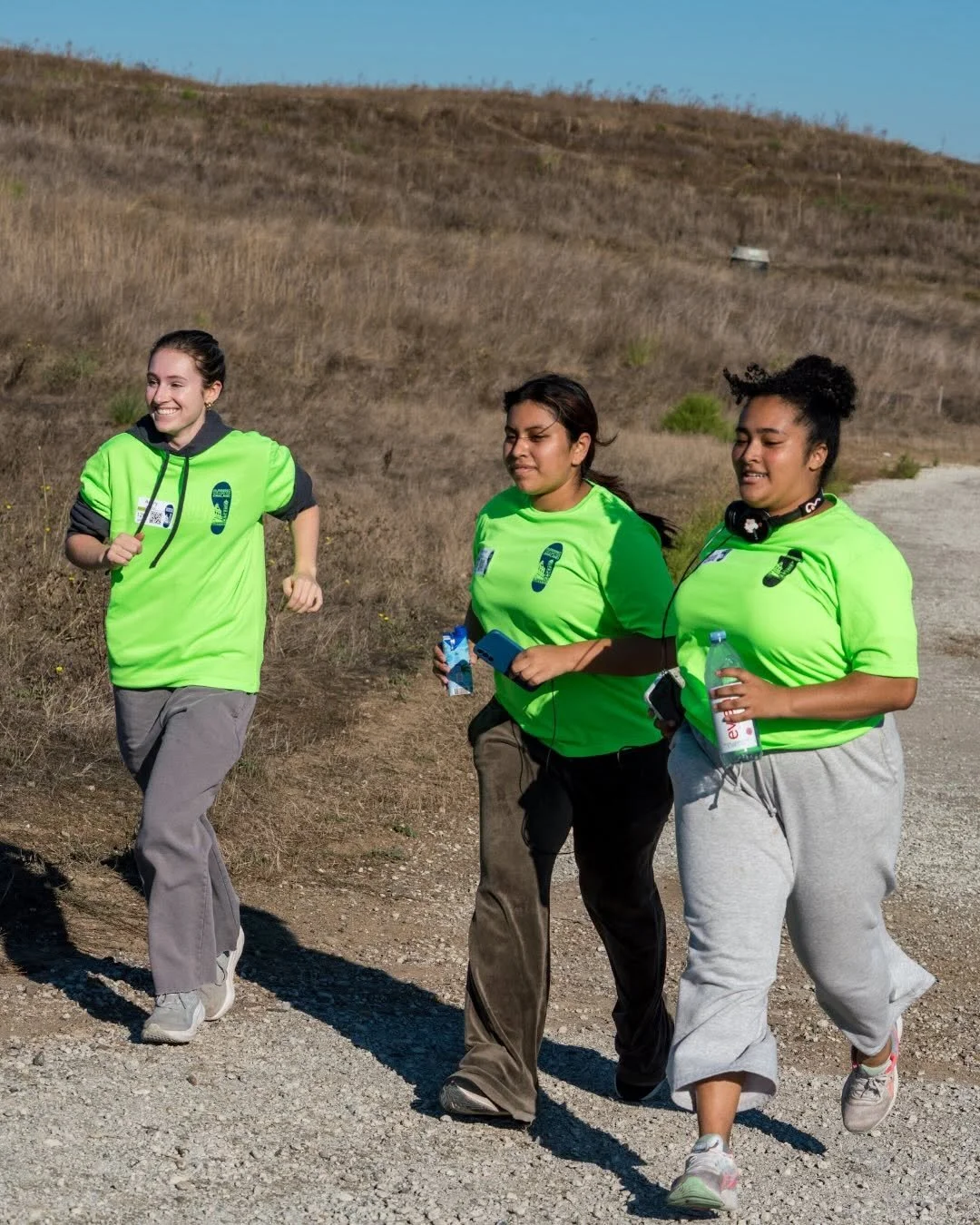 Three women running outdoors in a rural, hilly area, wearing bright green shirts and gray or brown pants, with one holding a water bottle and another holding a snack and a mobile phone.