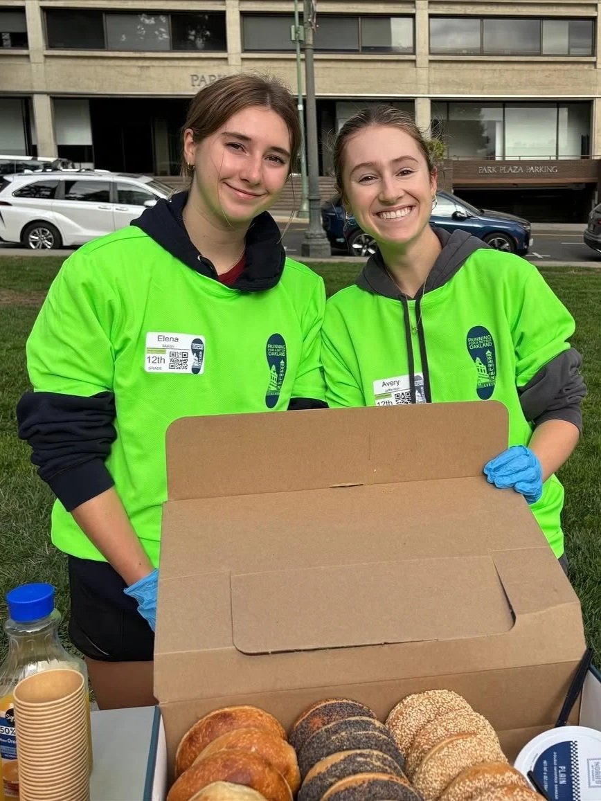 Two young women in bright green shirts standing outdoors behind a table with a box of bagels, smiling at the camera.