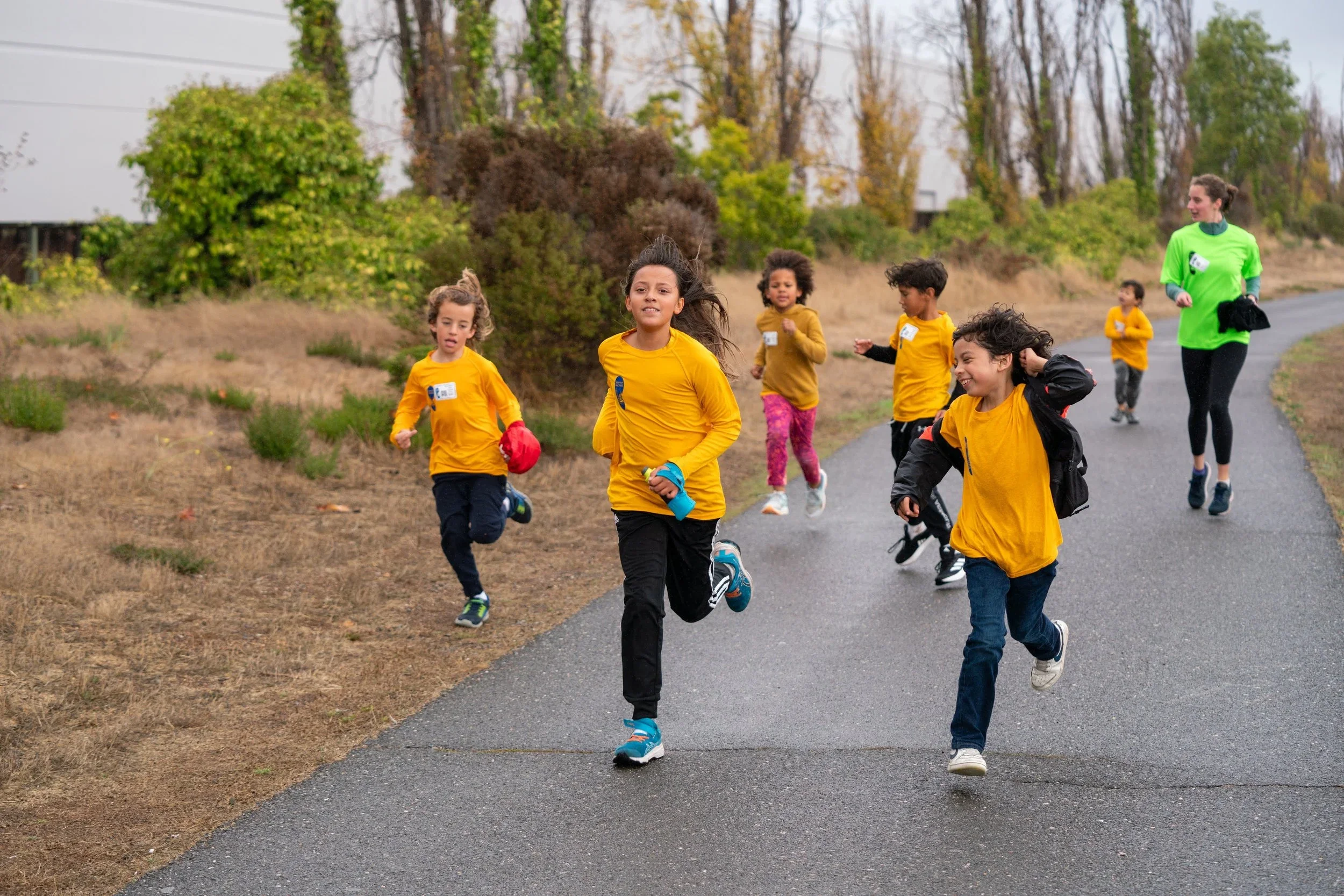 Children in yellow shirts running on a paved trail during a group outdoor activity, with a woman in a bright green shirt leading them, in a park-like setting with trees and grass in the background.