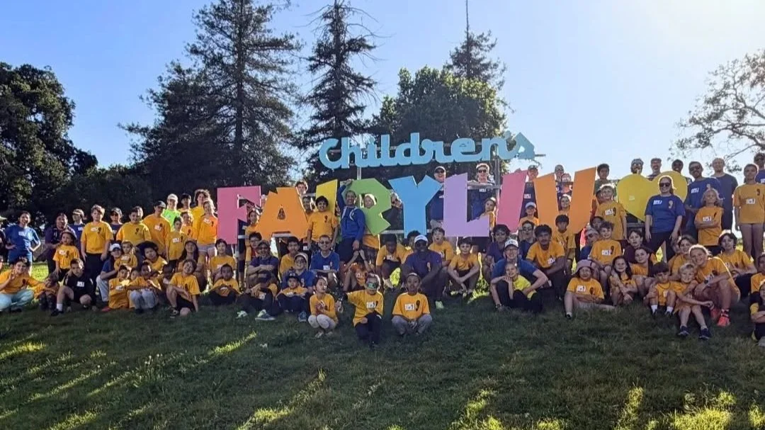 Group of children and adults gathered outdoors on a grassy hill, holding colorful large letters spelling 'Children's Family.' Trees and a clear sky are in the background.