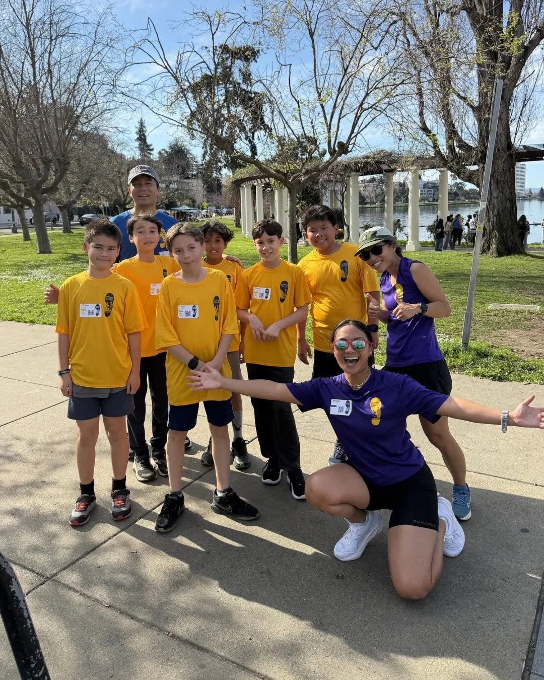 Group of children and adults in yellow and purple shirts posing outdoors in a park near a lake, smiling and making fun gestures on a sunny day.