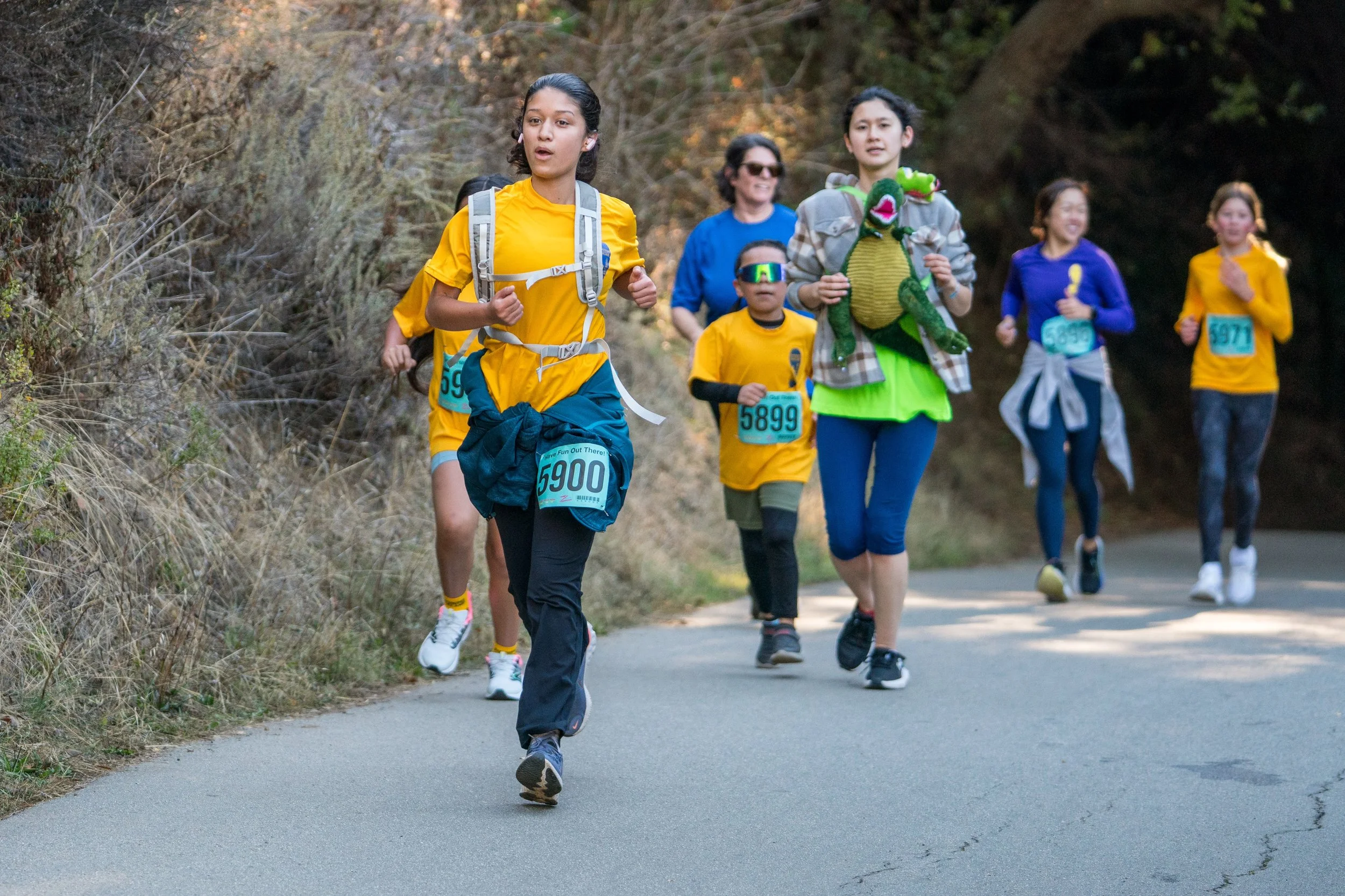 Un grupo de personas corre por un sendero en un parque a la luz del día. Llevan ropa deportiva de colores vivos y algunas tienen dorsales, lo que indica que participan en una carrera o en una carrera popular. Una de ellas lleva un disfraz de dragón.
