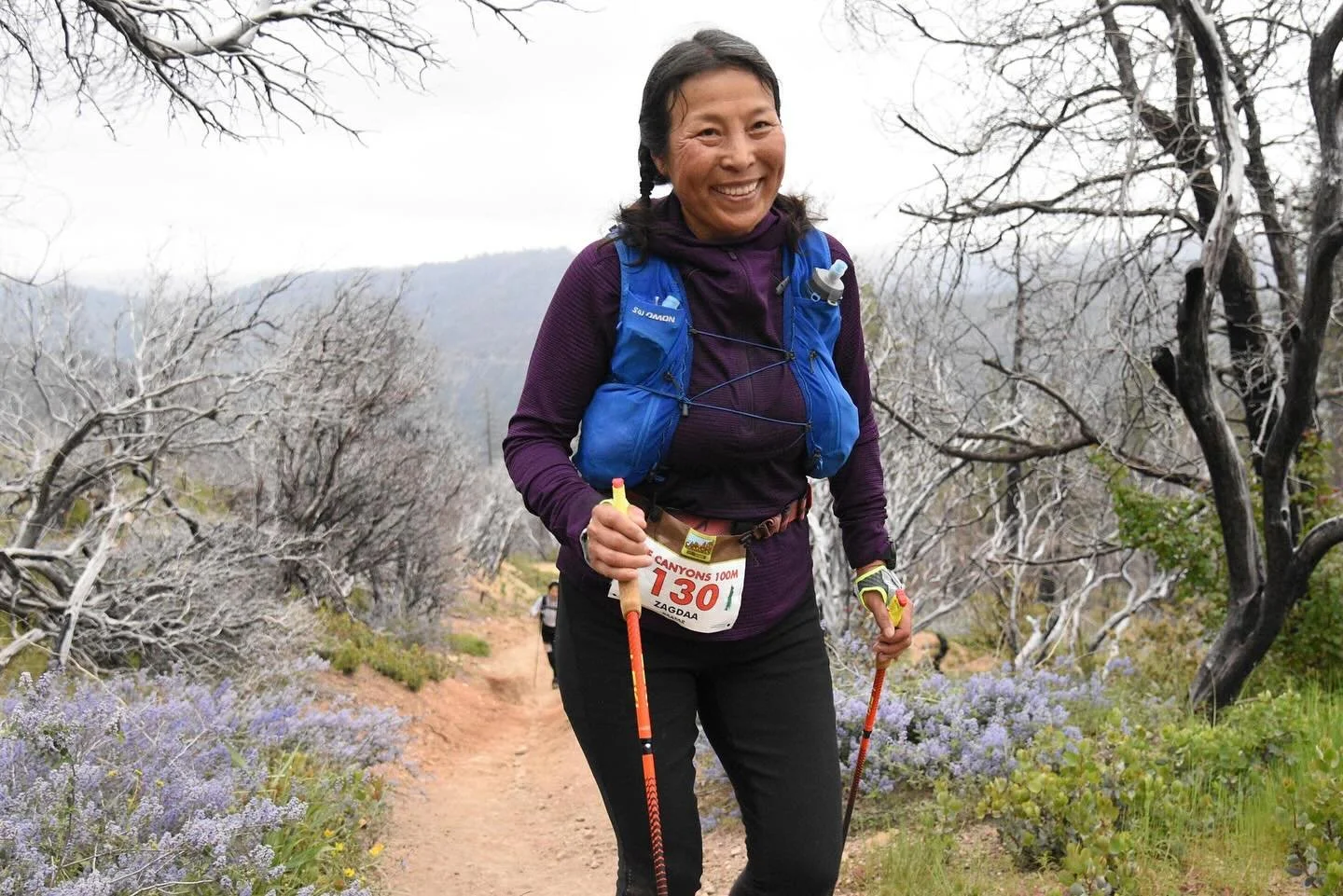 A woman smiling on a trail in a mountainous area, holding trekking poles, dressed in outdoor gear with a blue backpack, numbered bib, and surrounded by leafless trees and purple wildflowers.