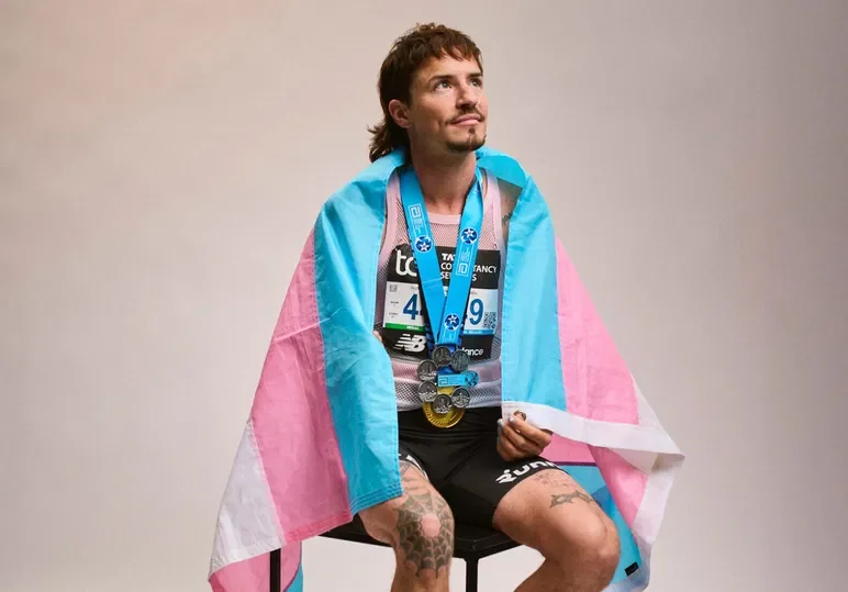 A male marathon runner with medals around his neck, sitting on a stool, draped in a transgender pride flag, holding a small item, and looking upward with a thoughtful expression.