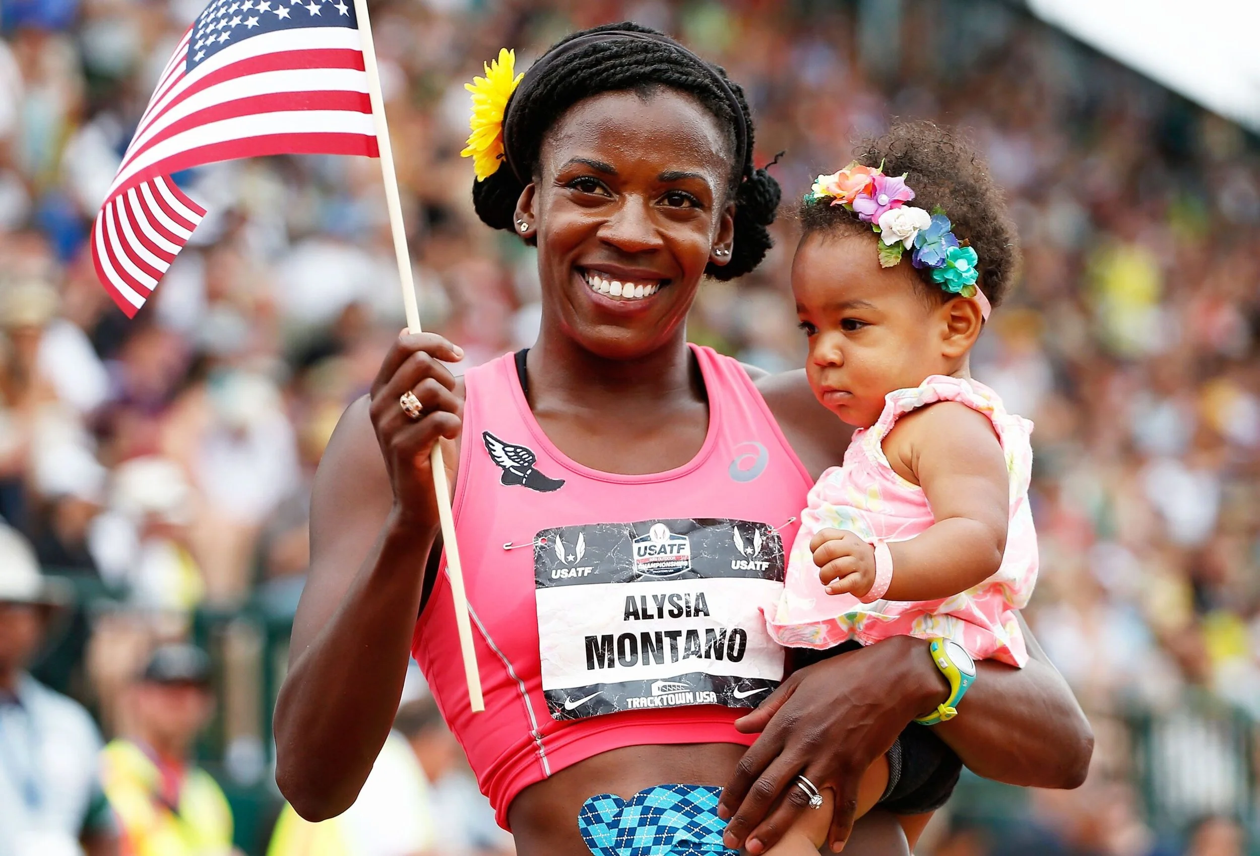 A woman in pink athletic gear holding a young girl with a colorful flower headband in a crowded outdoor event, woman waving an American flag.