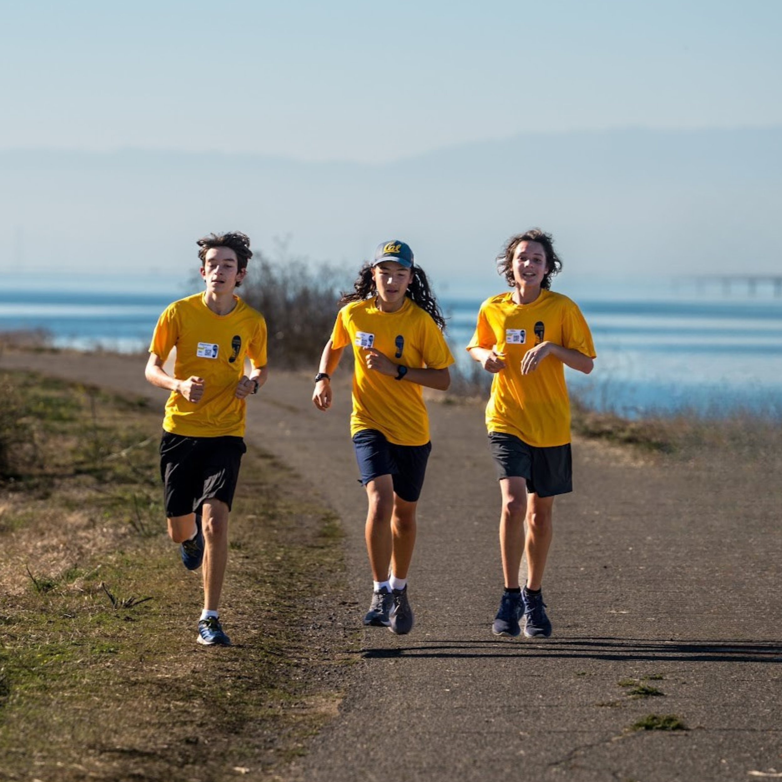 Three young runners in yellow shirts participating in an outdoor race near a body of water with a clear sky.