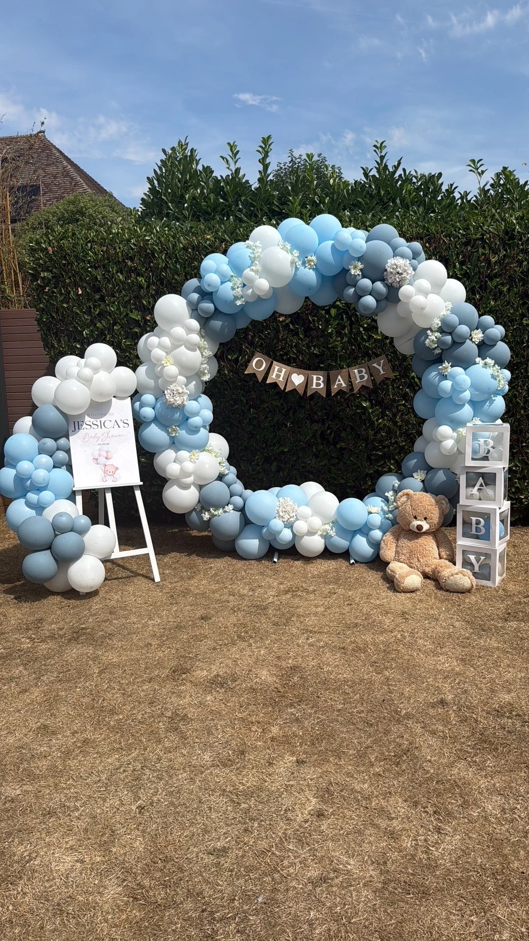 Baby shower decoration with a large blue, white, and gray balloon arch, teddy bear, sign that says "It's a Boy," and blocks spelling "Baby."