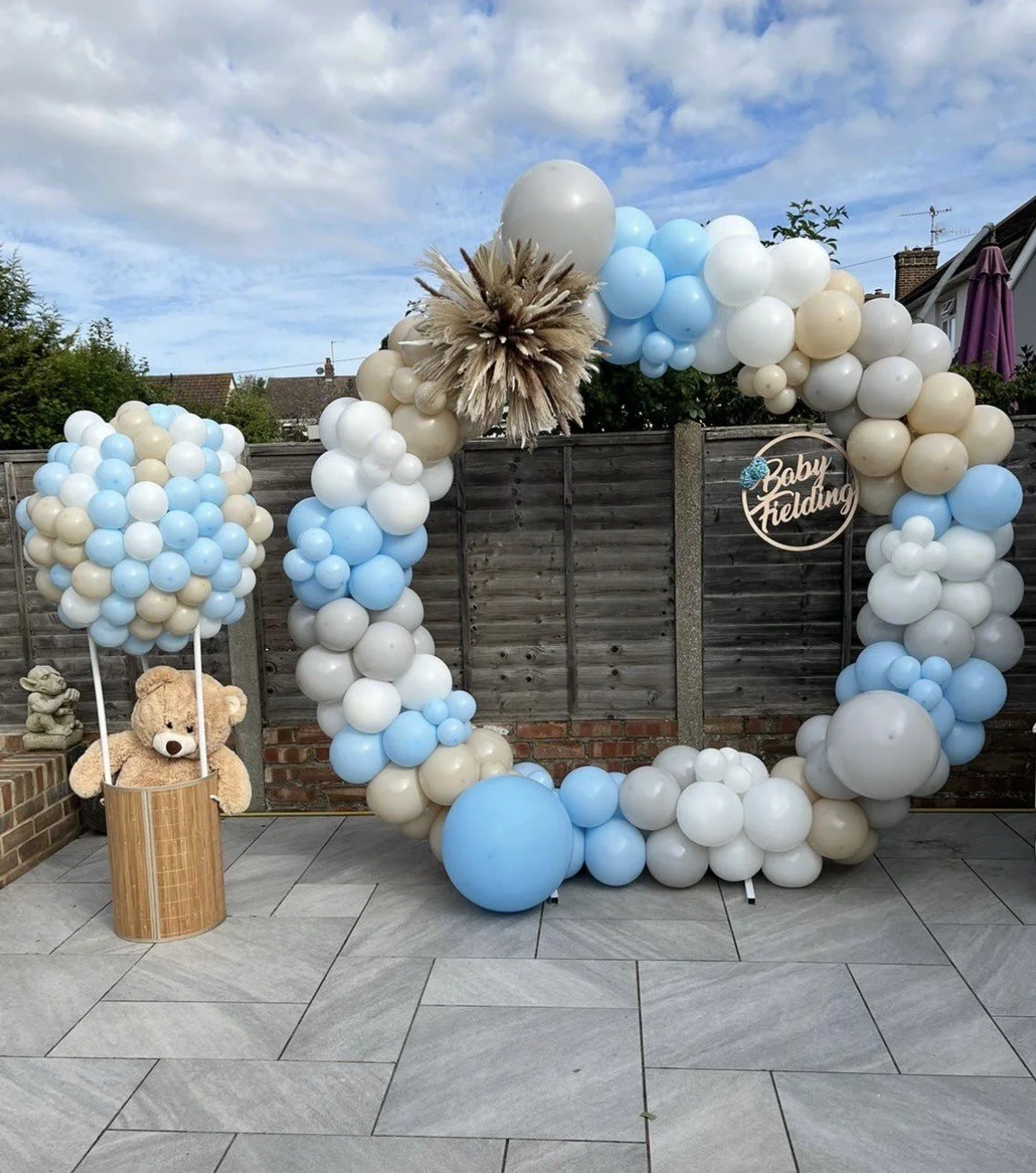 Balloon arch with blue, white, and beige balloons, a circular sign reading "Baby Felding," a teddy bear in a wooden basket, and cloudy sky in the background.