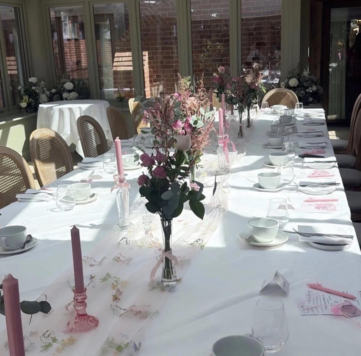 A long dining table decorated with pink candles, floral arrangements, and place settings for a special event in a sunlit room with large windows.