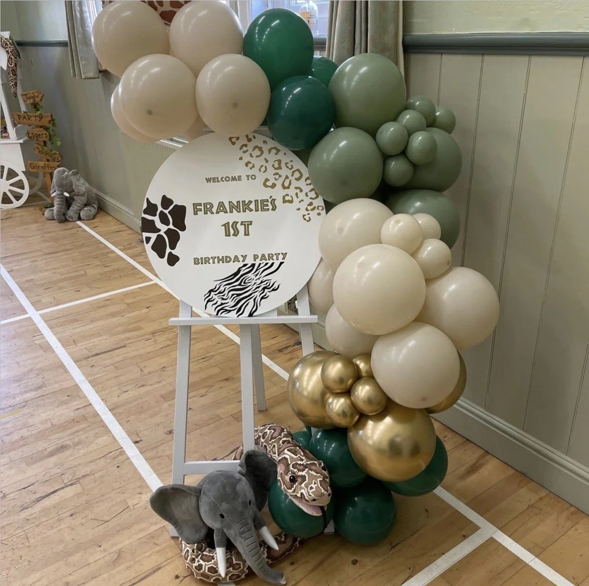 Balloon arrangement with shades of white, cream, green, and gold, and a sign that reads "Welcome to Frankie's 1st Birthday Party." There are plush elephant and giraffe toys on the floor next to the display.