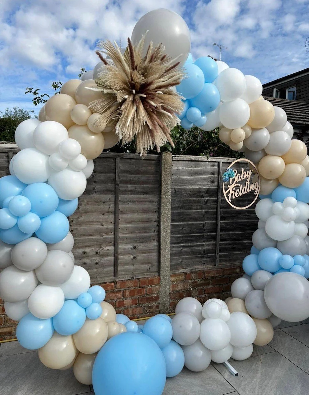 Balloon arch in shades of cream, white, and blue, with a pampas grass decoration at the top left, and a wooden circle sign reading 'Baby Felding' attached to the right side of the arch, outdoors against a wooden fence.