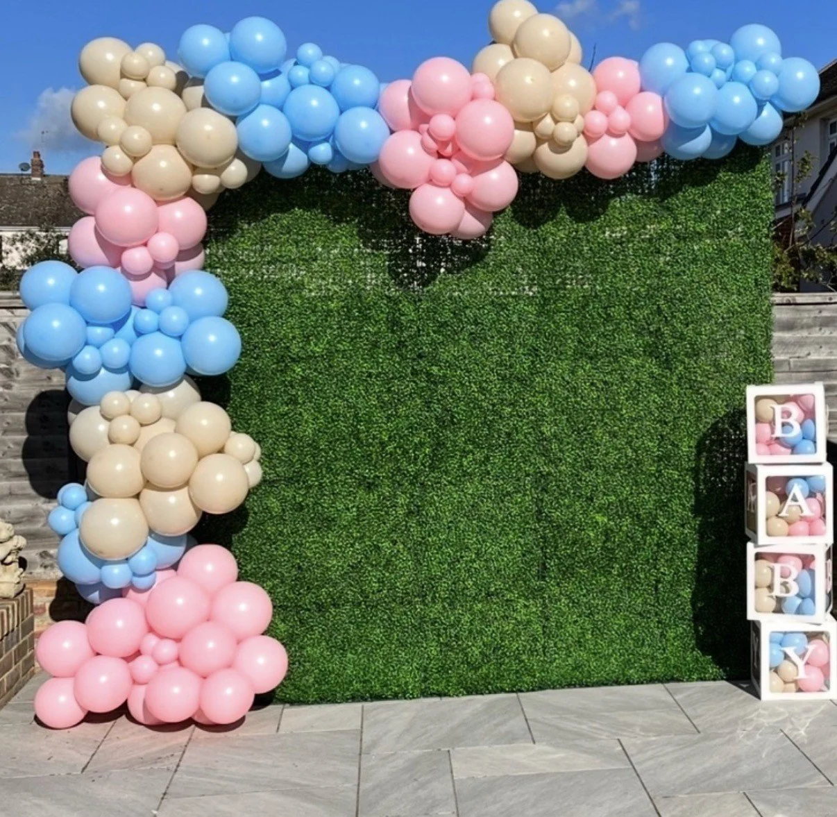 Decorative backdrop made of beige, pink, and blue balloons arranged around a green hedge wall. To the right, there are four white blocks with pink, blue, and beige balloons and letters spelling out 'BABY'.